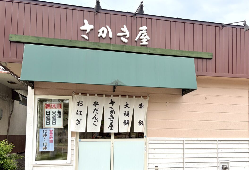 さかき屋製菓 有限会社さかき屋製菓 北海道 帯広市 西22条南2丁目21-7 郵便番号 080-2472 建物 外観 昭和レトロ 懐かしい佇まい 1階店舗 瓦屋根 木造建築 紺色の暖簾 白い文字 店舗看板 道路沿い 自立看板 縦長 老舗 お菓子屋 和菓子店 閉店の案内 告知の張り紙 店頭 ガラス戸 感謝のメッセージ 2026年7月31日 金曜日 営業終了 最終日 看板商品 串団子 みたらし団子 あんこ団子 醤油だれ 艶やか 焦げ目 焼き目 香ばしい もちもち 伝統の味 手作り 地域密着 コミュニティ（郵便番号080-2472、北海道帯広市西22条南2丁目21-7に位置する、地域密着の老舗和菓子店「有限会社さかき屋製菓」の昭和レトロでどこか懐かしい木造の建物外観。店舗正面の入り口には、店名が白い文字でくっきりと染め抜かれた格式ある紺色の暖簾が下がり、道路沿いには長年の歴史を感じさせる縦長の店舗看板が立てられている。店頭のガラス戸には、地域住民への深い感謝を伝えるために貼られた「2026年7月31日（金）の営業をもって閉店」と書かれた手書き風の閉店の案内（告知の張り紙）が掲示されている。手前の白い陶器の皿には、お店を代表する看板商品である手作りの串団子が綺麗に並んでおり、透明感のある艶やかな濃い茶色の醤油だれがたっぷりとかかり、直火で香ばしく炙られた均一な焦げ目（焼き目）がついた、弾力のあるもちもちとしたみたらし団子のシズル感あふれる質感が極めて詳細に写し出されている。）