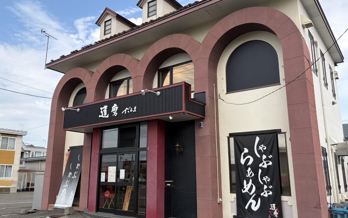 郵便番号080-2470、北海道帯広市西20条南4丁目44-19に所在する、しゃぶしゃぶ食べ放題とラーメンの和食チェーン・飲食店「食彩本舗 達磨（だるま）」の店舗建物とその敷地全体を捉えた屋外の外観写真。平屋建ての商業用建物の正面エントランス側が写っており、店舗の壁面や屋根付近、独立したロードサイド看板には「食彩本舗 達磨」という店名ロゴの文字がはっきりと確認できる。手前側にはアスファルトで舗装された広い駐車場のスペースが広がっており、車止めブロックや白線、敷地境界の道路が写り込んでいる。日中の明るい時間帯に撮影された、店舗の営業風景と周囲の立地環境が客観的に伝わる全景写真。